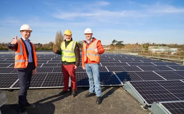 Three men in hard hats and high-visibility jackets standing among rows of solar panels on the roof of Woodside Leisure Centre in Watford