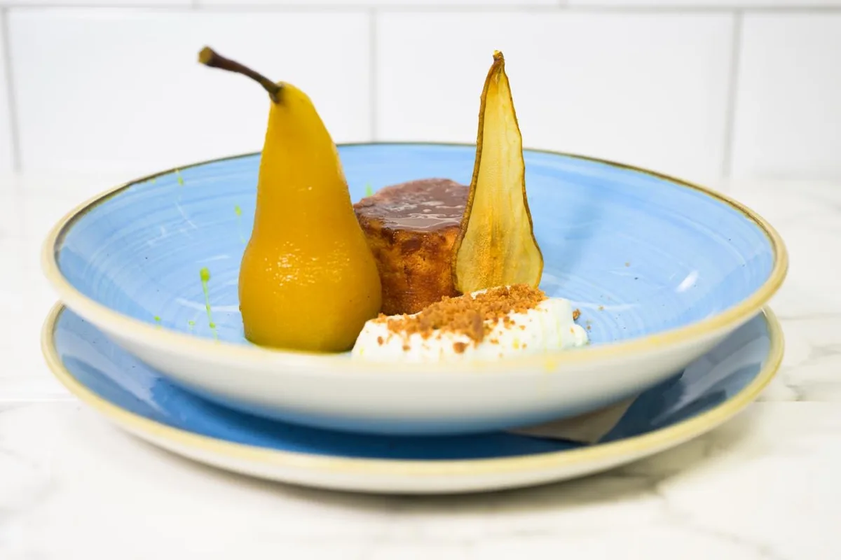 A blue plate with a whole poached pear, a round ginger cake glazed in sauce, a quenelle of baked yoghurt with crumb on top, and a thin pear crisp standing upright